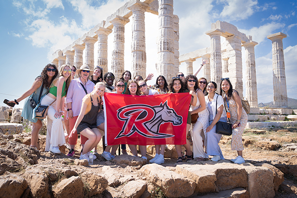 A group of students studying abroad stand in front of Greek ruins holding a Rider flag