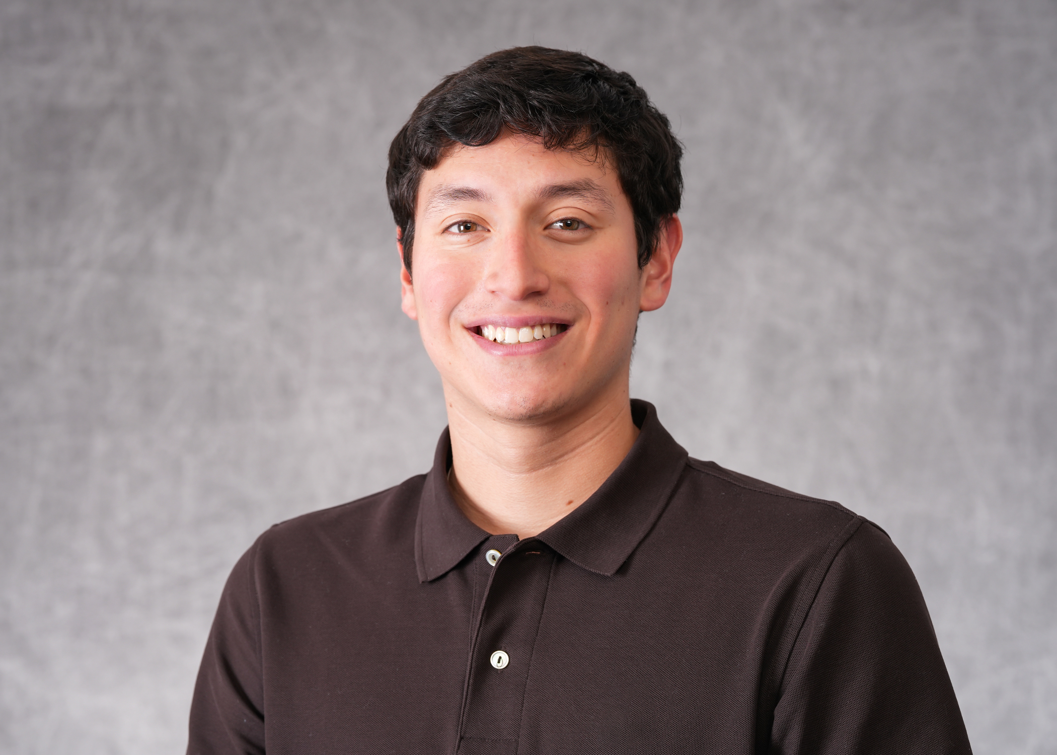 Male student headshot in front of a gray background. The student has dark, short hair and is wearing a brown polo shirt. 