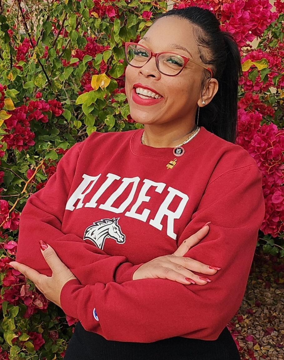 Female student wearing a red Rider University sweatshirt poses in front of rose bushes. The Student is wearing Bright red glasses and has dark hair pulled into a ponytail. 