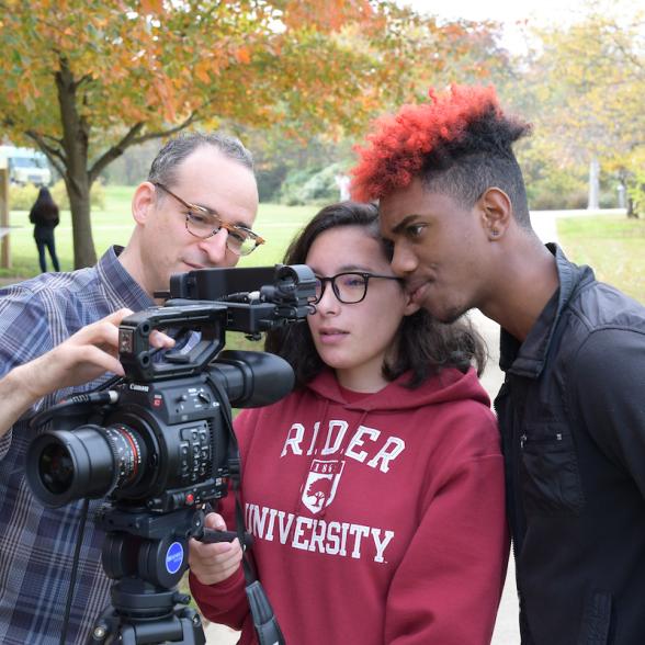 Two students and a professor looking through a large camera lens 