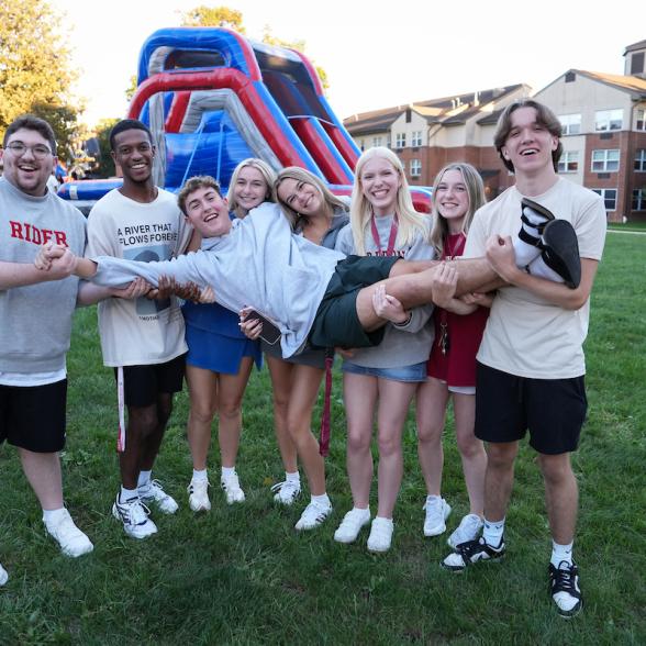 Eight Rider University students smile and pose together on the Campus Green, holding one student horizontally, with a colorful inflatable slide and campus residence halls in the background.