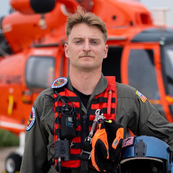 Coast Guard officer poses in front of orange rescue helicopter. He's dressed in tactical gear and holding his helmet. 