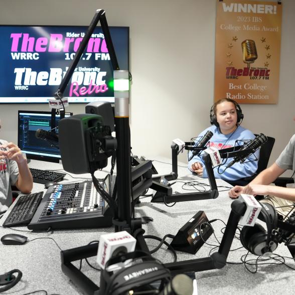 Three students sit a radio command center table. There is a lot of equipment present. 