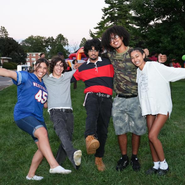 Five Rider University students smile and pose with their arms around one another on the Campus Green during an outdoor celebration, with games and inflatable attractions in the background.
