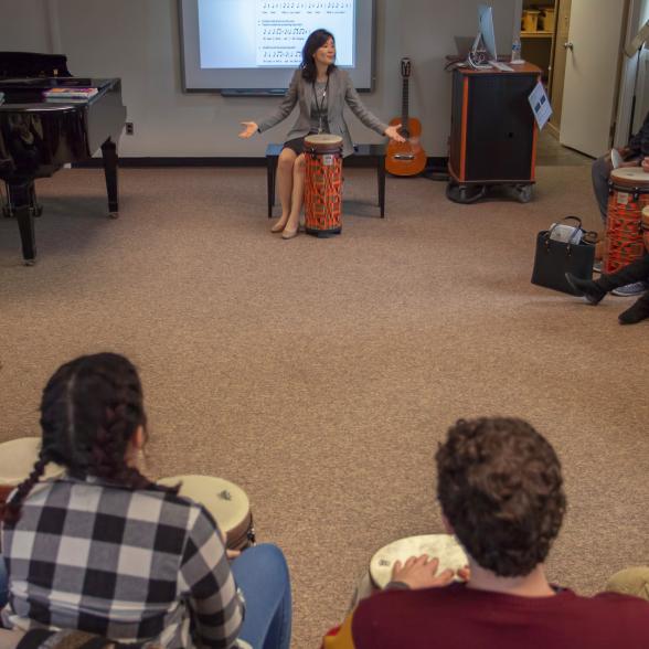 Music educator sits in a circle of students with bongos giving a lecture