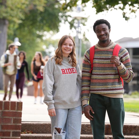 Two Rider students on Rider campus facing the camera and smiling, one is wearing a Rider sweatshirt and the other a striped sweater. There are students laughing in the background
