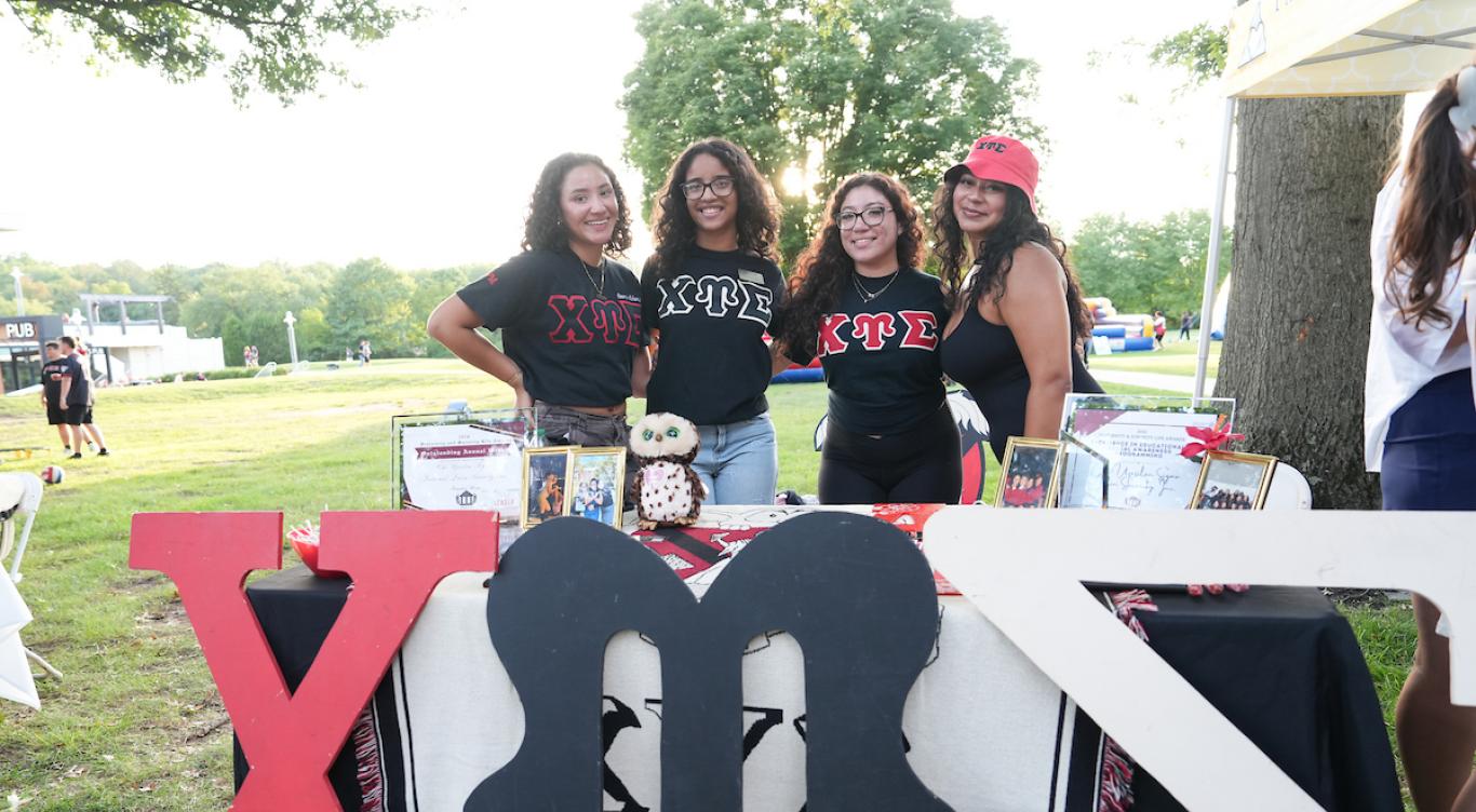 Four members of Chi Upsilon Sigma pose together behind their sorority’s display table at an outdoor campus event. The table features framed photographs, awards, Greek-letter memorabilia, and a plush owl.