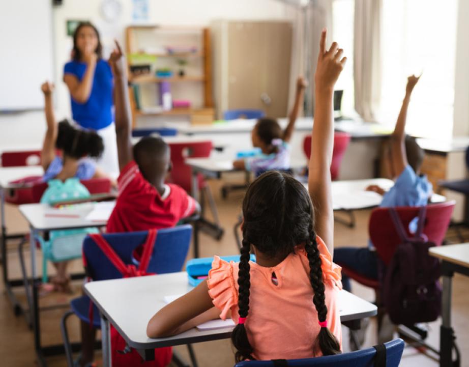 Students in elementary classroom raise hands in response to teacher's lesson
