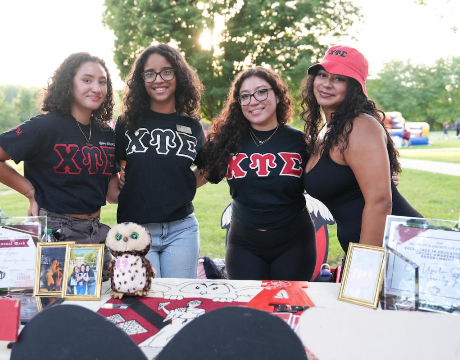 Four members of Chi Upsilon Sigma pose together behind their sorority’s display table at an outdoor campus event. The table features framed photographs, awards, Greek-letter memorabilia, and a plush owl.