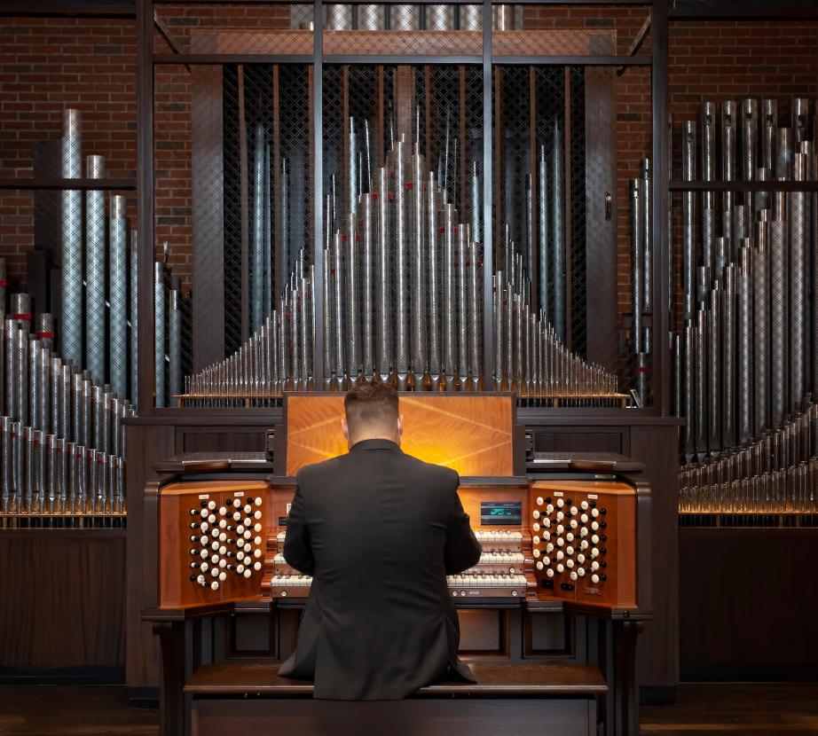 A man has his back to the camera as he sits at the organ, surrounded by pipes in Gil Chapel