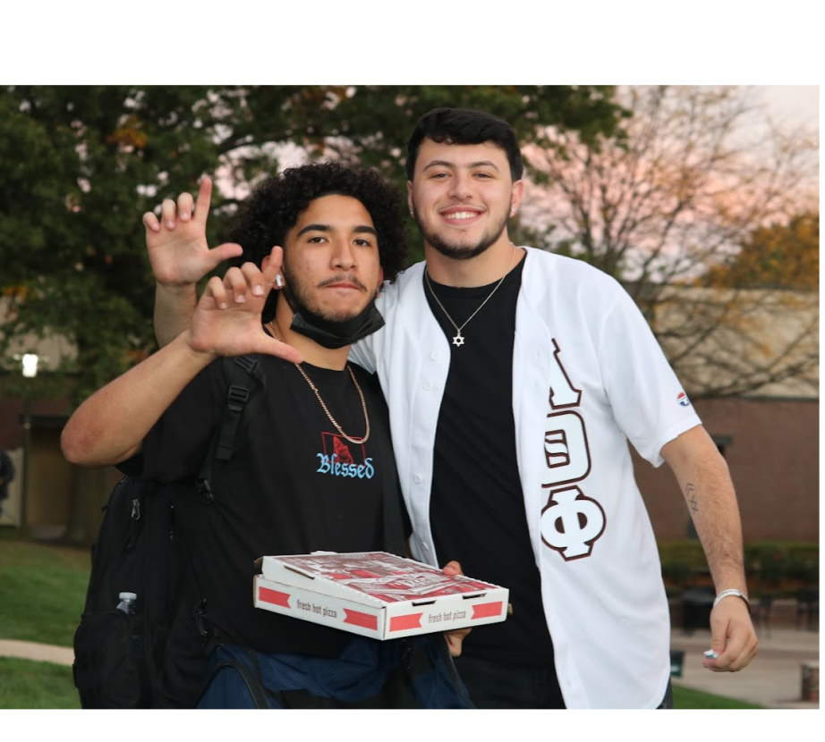 Two Multicultural Greek Council students hold up hand signs while holding pizza box