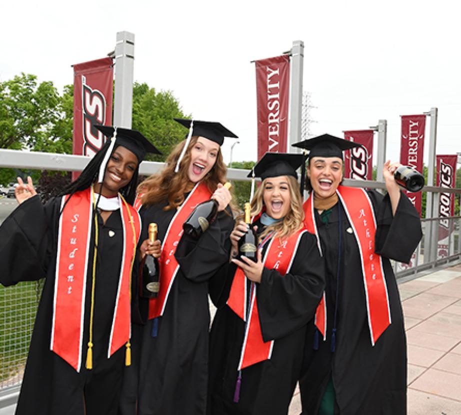 Group of graduates pose on the Rider campus with their caps and gowns, holding bottles of champagne