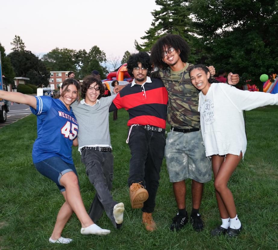 Five Rider University students smile and pose with their arms around one another on the Campus Green during an outdoor celebration, with games and inflatable attractions in the background.
