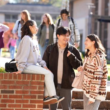 Three students sit outside talking. One student is sittong on a low wall's ledge, the other two are on the steps next to the wall.